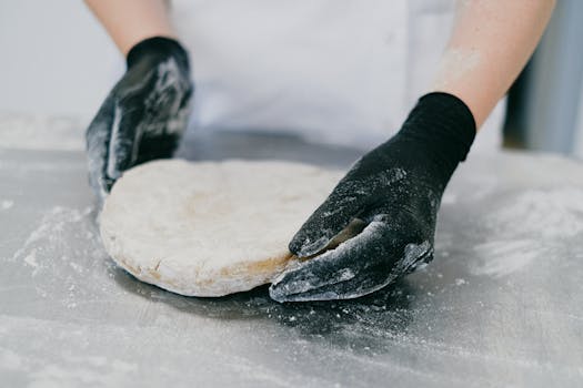 pexels-photo-5964530-5964530 Close-up of a chef with black gloves kneading dough on a silver surface, showcasing bakery skills.