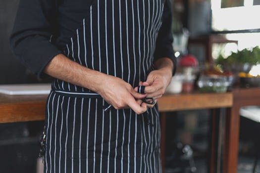 pexels-photo-8477052-8477052 Chef tying a black and white striped apron in an indoor kitchen with fresh ingredients nearby.